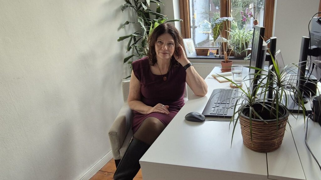 Die Mediendesignerin Barbara Behrens sitzt in ihrem Arbeitszimmer mit vielen Pflanzen und blickt in die Kamera.